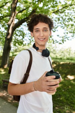 Kıvırcık saçlı genç bir adam güneşli parkın tadını çıkarırken ılık bir içecek yudumluyor..