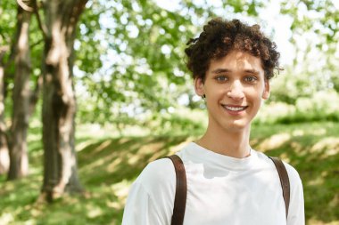 Kıvırcık saçlı genç bir adam ağaçlarla dolu güneşli yeşil bir parkta dinlenirken sıcacık gülümsüyor..