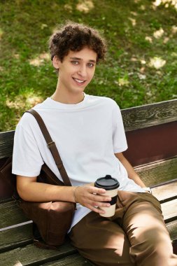 Beyaz tişörtlü genç bir adam yemyeşil bir parktaki bankta otururken kahvesinin tadını çıkarıyor..