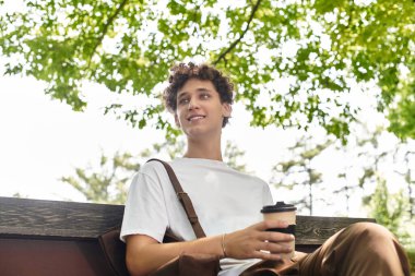 Genç bir adam yeşil bir parkta oturur, güneşli havanın tadını çıkarırken bir içki yudumlar..