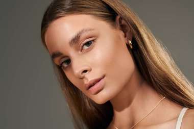 Genç bir kadın, bronz tenini ve doğal makyajını gri arka plan yaz titreşimleriyle ön plana çıkarıyor.