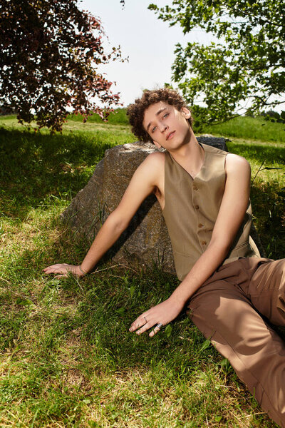A stylish young man with curly hair relaxes on a rock in a lush green park, embracing summer vibes.