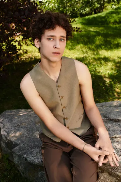 Kıvırcık saçlı genç bir adam, parkta şık bir yaz kıyafeti giymiş bir taşın üzerinde dinleniyor..