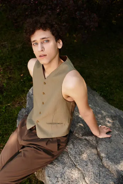 Kıvırcık saçlı şık bir genç adam bir kayanın üzerinde dinleniyor, yaz güneşini parkta emiyor..