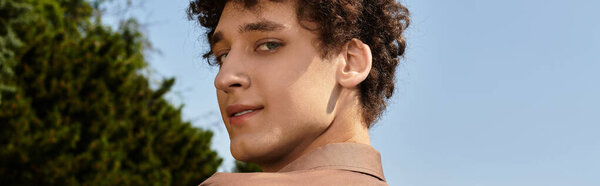 A young man with curly hair relaxes in a lush park, under the sun, banner