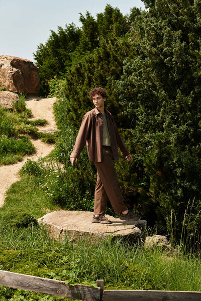 Curly-haired man in fashionable summer attire walks along a park's natural path.