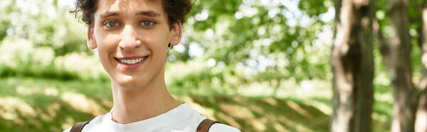 A young man with curly hair smiles while relaxing in a sunny park surrounded by greenery, banner
