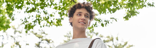 A young man with curly hair smiles while spending time outdoors in a vibrant summer park, banner