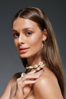 Kendine güvenen genç bir kadın, stüdyo ortamında deniz kabuğu kolyesiyle yaz stili sergiliyor..