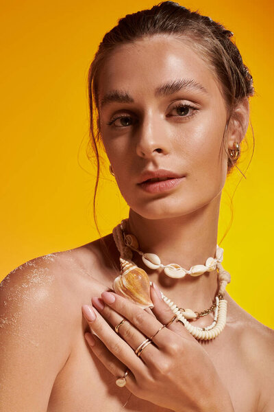 A beautiful young woman poses confidently, highlighting her seashell jewelry in a warm studio.