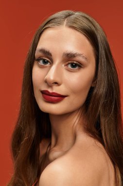 Genç bir kadın zarif bir şekilde canlı turuncu bir arka plana poz veriyor, güzelliğini vurguluyor..