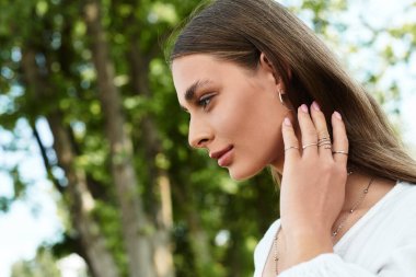 Büyüleyici, uzun saçlı genç bir kadın. Yeşilliklerin arasında hafifçe mücevherlerine dokunuyor..