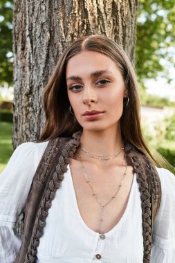 Gün ışığı yaprakların arasında dans eder. Genç ve güzel bir kadın güvenli bir şekilde bir ağacın yanında poz verir..