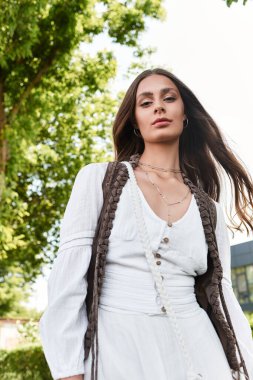 Şık giyinmiş kendine güvenen genç bir kadın güneşli, canlı bir parkta duruyor..