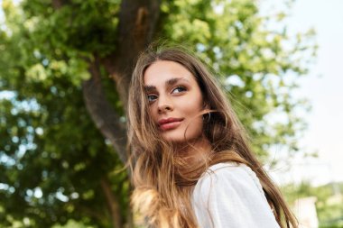 Güzel genç bir kadın yavaşça gülümsüyor, canlı ağaçların arasında güneşlenerek..