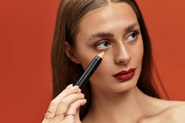 Genç ve güzel bir kadın canlı bir arkaplanda poz verirken kalemle makyaj yapıyor..