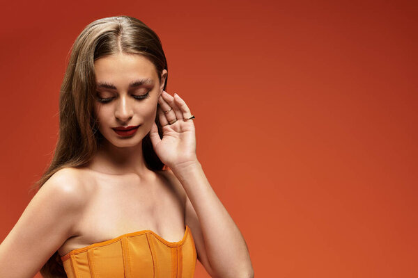 Молодая женщина с длинными волосами элегантно позирует, прижав руку к лицу, в стильном топе.