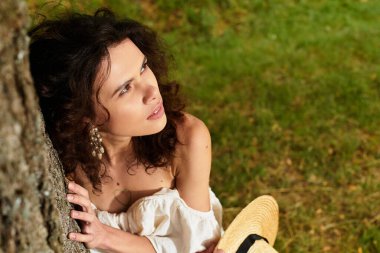 Beyaz yazlık elbiseli kıvırcık saçlı kadın huzur parkındaki bir ağaca yaslanıyor..