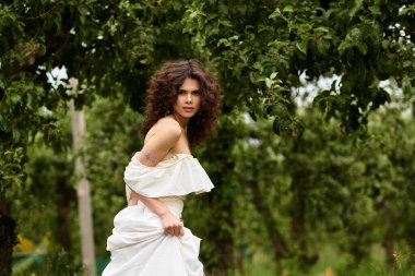 Kıvırcık saçlı hoş bir kadın yeşil bir parkta dolanıyor, yazın cazibesini kucaklıyor..