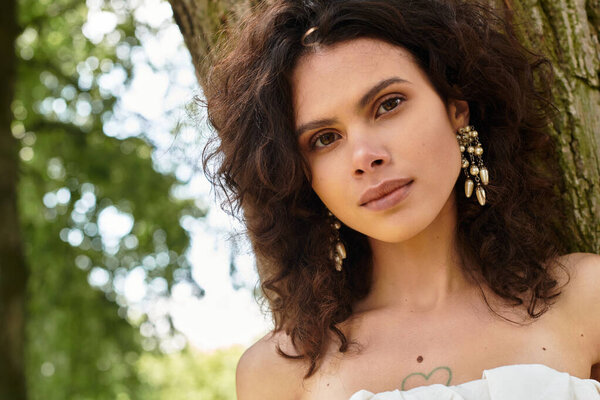 Young woman with curly hair and earrings captivates in a sunlit park surrounded by greenery.
