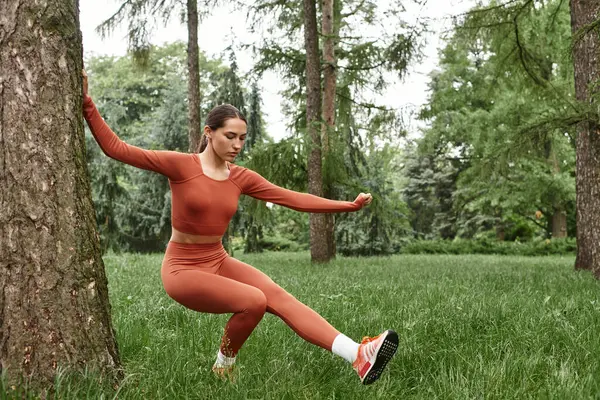 Genç bir kadın, yemyeşil bir ortamda, bir ağacın yanında yoga yapıyor, huzur ve refahı teşvik ediyor..