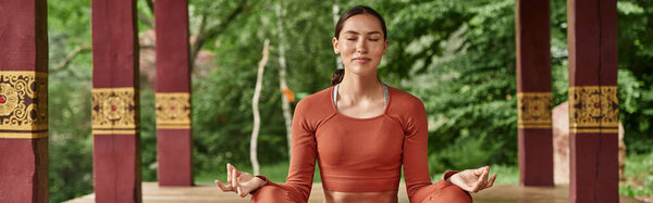 A young beautiful woman teaches yoga while meditating peacefully in a lush outdoor space.