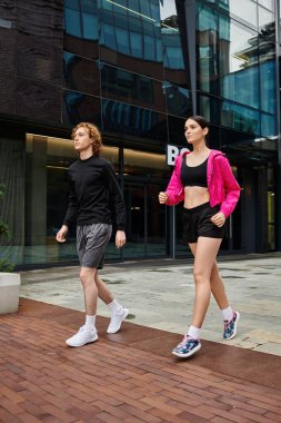 İki genç sporcu enerjik bir kıyafetle modern bir şehirde yürüyor..