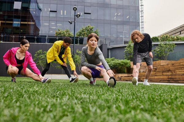 Four young athletes warm up by stretching in a green urban park with modern buildings nearby.