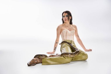 Genç bir kadın beyaz bir zeminde poz veriyor, bacaklarının etrafında bir yılan, güzelliği ve tehlikeyi harmanlıyor..