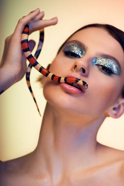 Kendine güvenen, sanatsal makyajlı genç bir kadın yüzünde renkli bir yılan tutuyor..