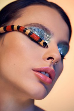 Çarpıcı makyajlı büyüleyici bir kadın renkli bir yılanla poz verip büyüleyici bir atmosfer yaratıyor..