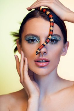 Büyüleyici kadın kendine güvenen pozlar veriyor. Yüzüne bir yılan sarılmış. Güzellik ve tehlikeyi harmanlıyor..