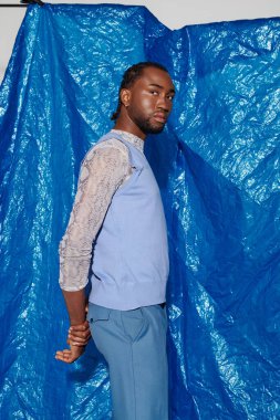 Şık bir genç adam parlak mavi bir arka plana karşı duruyor, cazibesini ve güvenini sergiliyor..