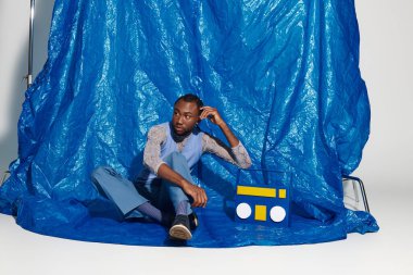 Şık bir genç adam, modern modayı gözler önüne seren mavi bir arkaplanda gelişigüzel oturuyor..