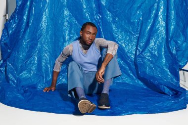 Şık giyinmiş genç bir adam göz kamaştırıcı bir şekilde ışıldarken cesur mavi bir arka planda poz verir..