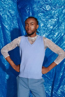 Şık bir genç adam kendine güvenerek ayakta duruyor, elleri kalçasında, mavi kıyafetli bir cazibe yayıyor..