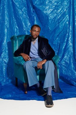 Şık bir genç adam mavi arka planlı bir sandalyede rahatlar..