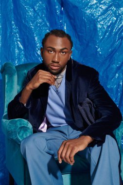 Genç adam kendine güvenerek bir sandalyeye oturur, mavi bir zemine karşı şık kıyafetlerini sergiler..