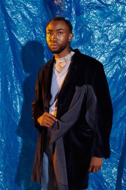 Şık giyinmiş genç bir adam dimdik duruyor, koyu mavi renkler ve büyüleyici bir görünüş ile çerçevelenmiş..