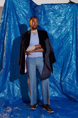 Şık giyinmiş yakışıklı bir adam dinamik mavi bir arkaplan önünde duruşuyla poz verir..