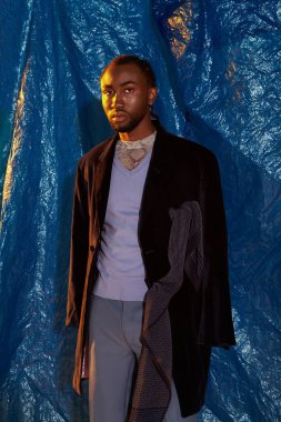 Zarif giyinmiş şık bir genç adam çarpıcı bir mavi arkaplanın ortasında duruyor, büyüleyici bir cazibe ve duruş..