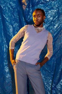 Şık giyinmiş kendine güvenen genç bir adam cesur mavi bir fon önünde poz verir..