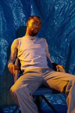 Yakışıklı genç adam rahat bir şekilde oturur, dikkat çekici mavi bir arka planla rahatça arkasını gösterir..