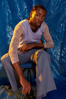 Yakışıklı genç adam canlı mavi tonlara karşı şık bir elbise içinde otururken düşünceli bir şekilde poz veriyor..