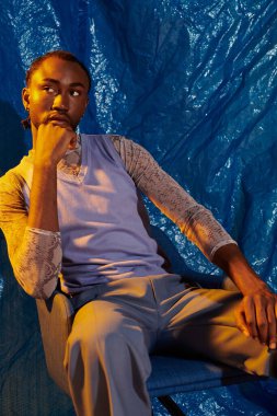 Yakışıklı bir genç adam düşünceli bir şekilde oturur, canlı mavi bir arka plan tarafından çerçevelenir..