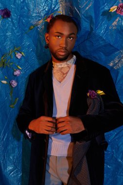 Yakışıklı genç bir adam koyu renk bir ceket ve açık renk süveter içinde dikiliyor, etrafı çiçeklerle çevrili..