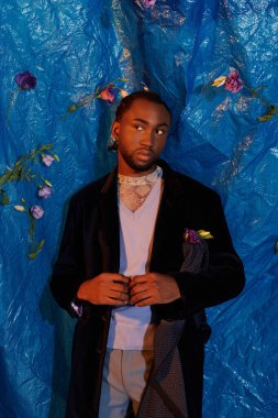 Şık bir genç adam mavi bir arka fonun önünde duruyor, zarafet ve çekicilik sergiliyor..