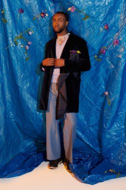 Şık bir genç adam çiçeklerle çevrili canlı bir mavi arkaplanın önünde özgüvenle duruyor..