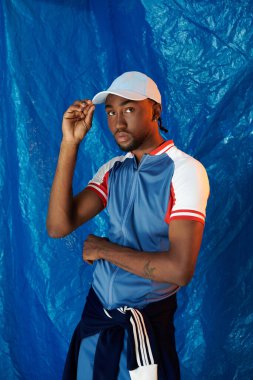 Yakışıklı genç adam spor kıyafetleri içinde mavi arka planda kendine güvenini gösteriyor..