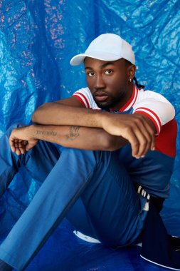 Yakışıklı genç bir adam düşünceli bir şekilde yere oturmuş mavi bir elbise giyiyor..